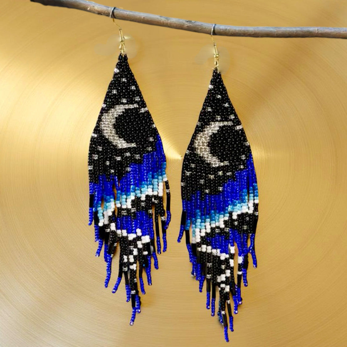Handmade beaded earrings with a crescent moon design at the top and long black, blue, and white beaded fringe, finished with hypoallergenic 316L surgical steel lever back hooks against a gold background