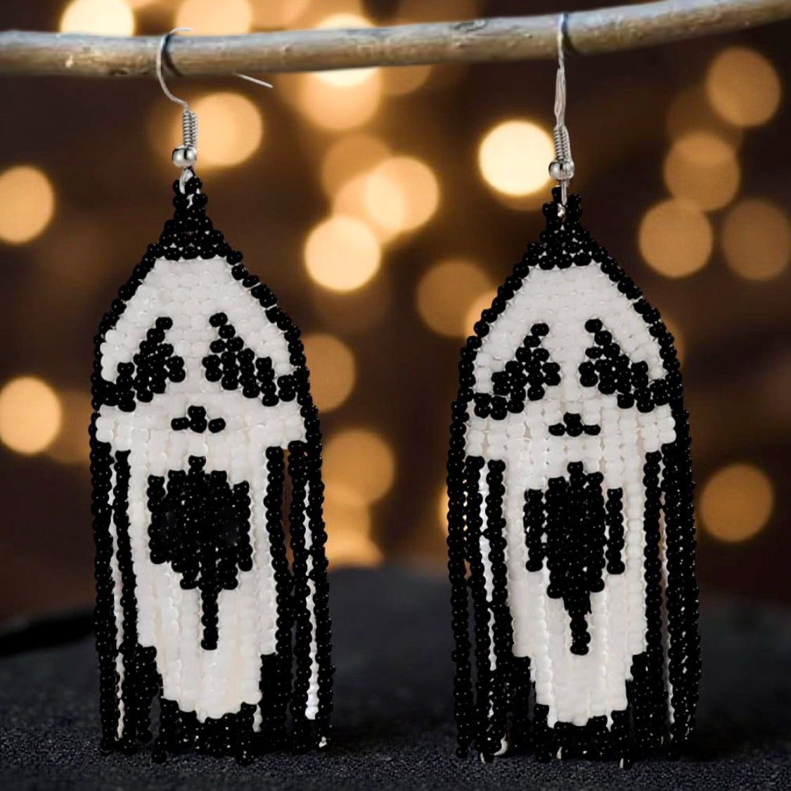 A pair of handmade beaded earrings featuring a black and white ghost face design in the fringe, hanging from hooks against a bokeh background.