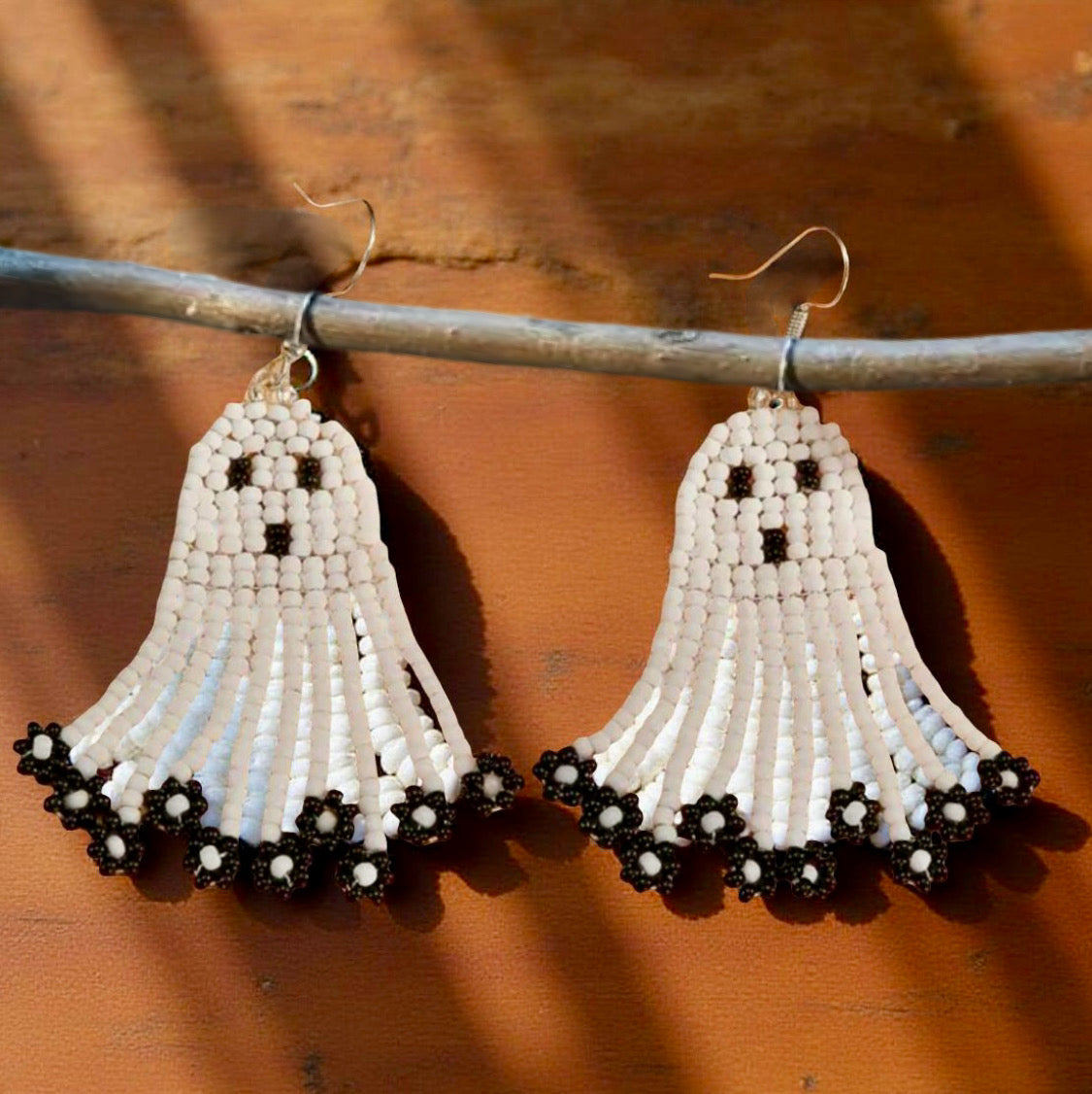 Handmade beaded earrings in the shape of cute white ghosts with black facial details and floral black fringe, perfect for Halloween and spooky season style. Wood background