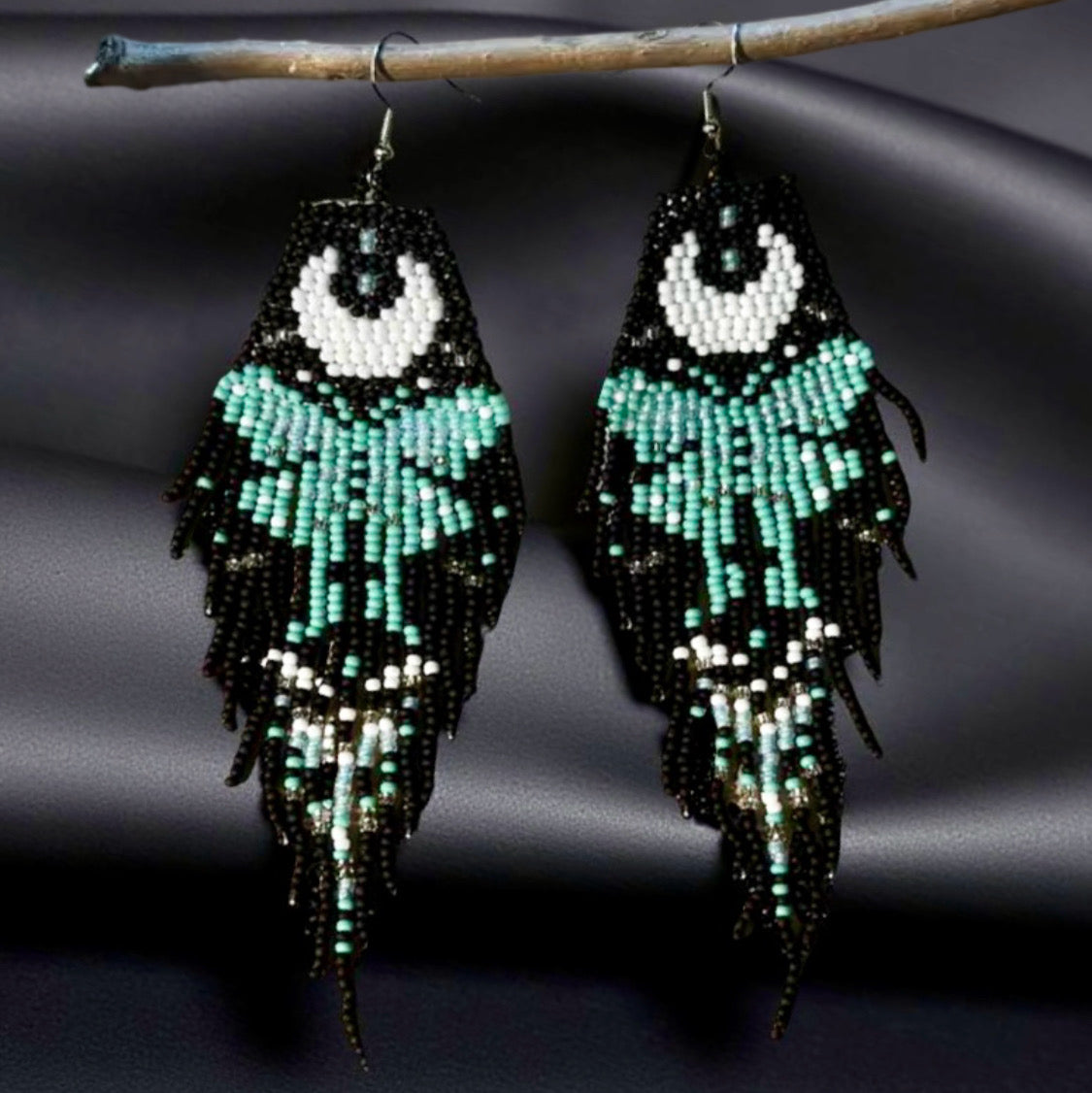 Handmade beaded earrings with a moon and turquoise butterfly design, featuring a black fringe. The earrings are hung from hypoallergenic 316L surgical steel lever backs. Black satin background