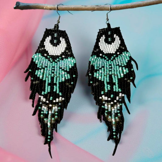 Handmade beaded earrings with a moon and turquoise butterfly design, featuring a black fringe. The earrings are hung from hypoallergenic 316L surgical steel lever backs. Pink and blue background