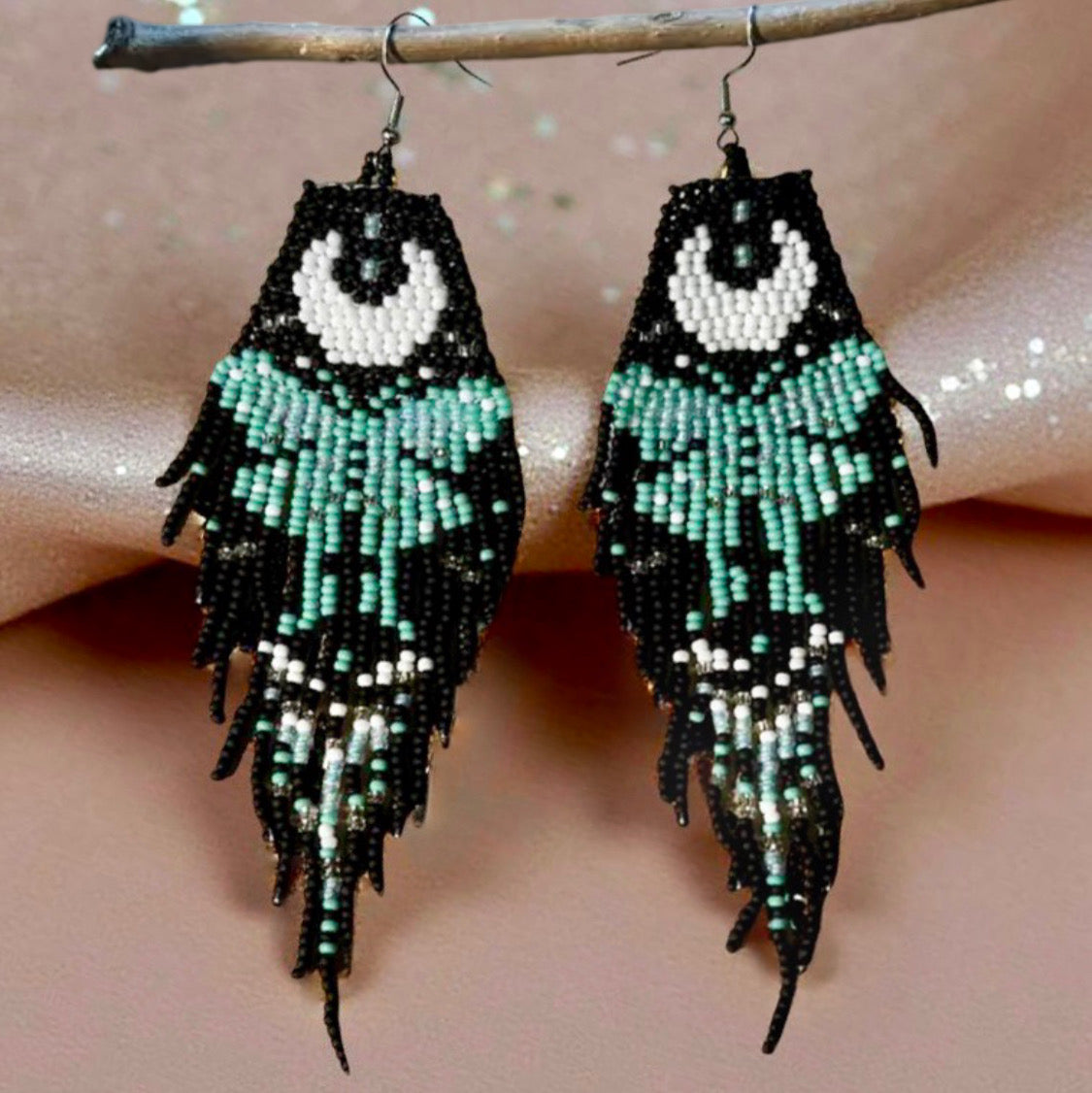 Handmade beaded earrings with a moon and turquoise butterfly design, featuring a black fringe. The earrings are hung from hypoallergenic 316L surgical steel lever backs. Shimmery fabric background