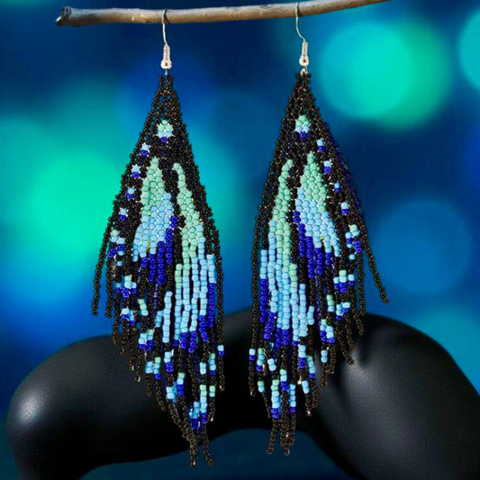 Handmade beaded earrings featuring a blue butterfly wing fringe, with intricate beadwork in shades of turquoise and blue. The earrings are attached to hypoallergenic 316L surgical steel lever backs. Black prop