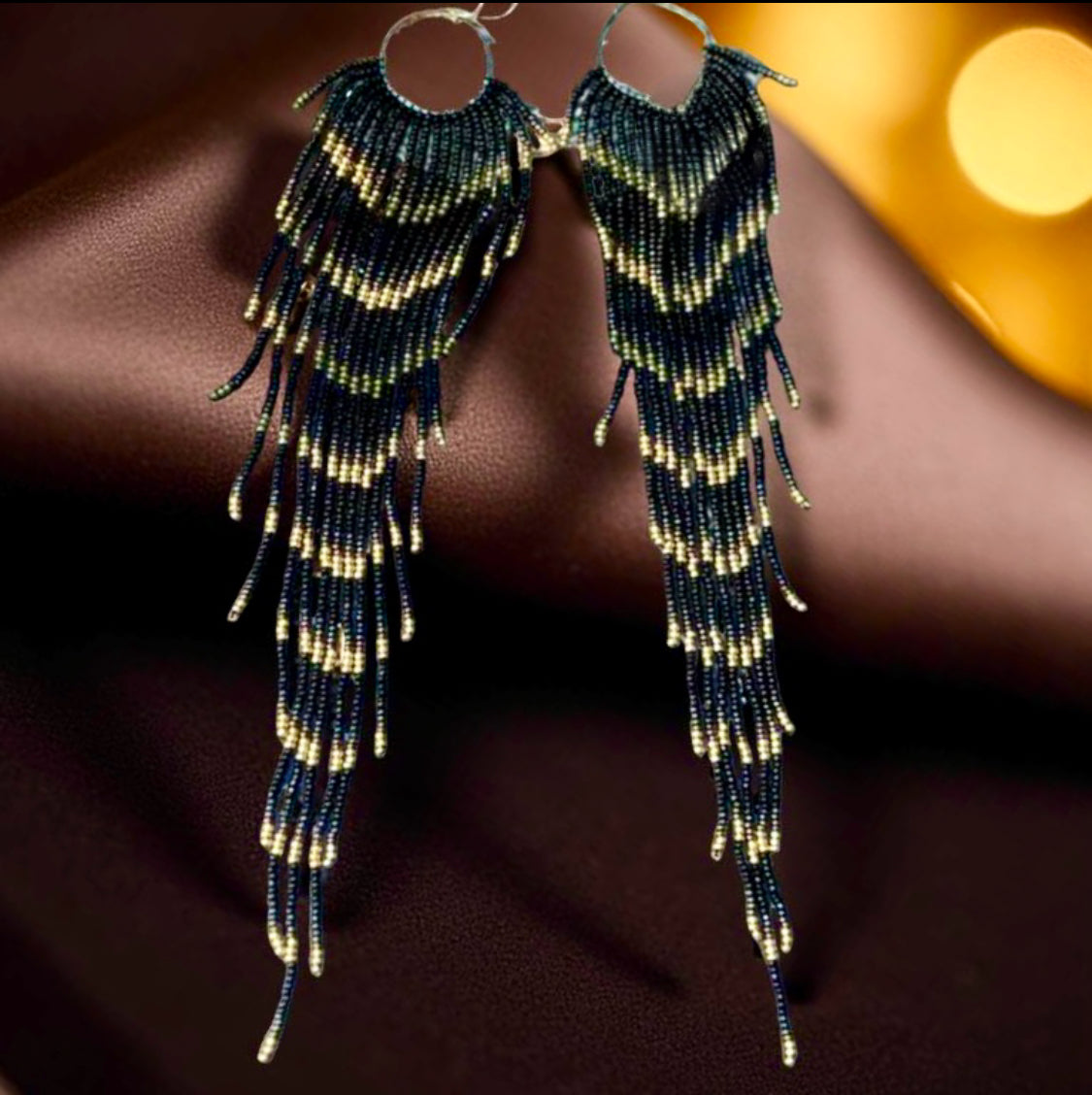 Handmade beaded earrings with extra-long black, green, and gold fringe displayed on a soft chocolate satin background.