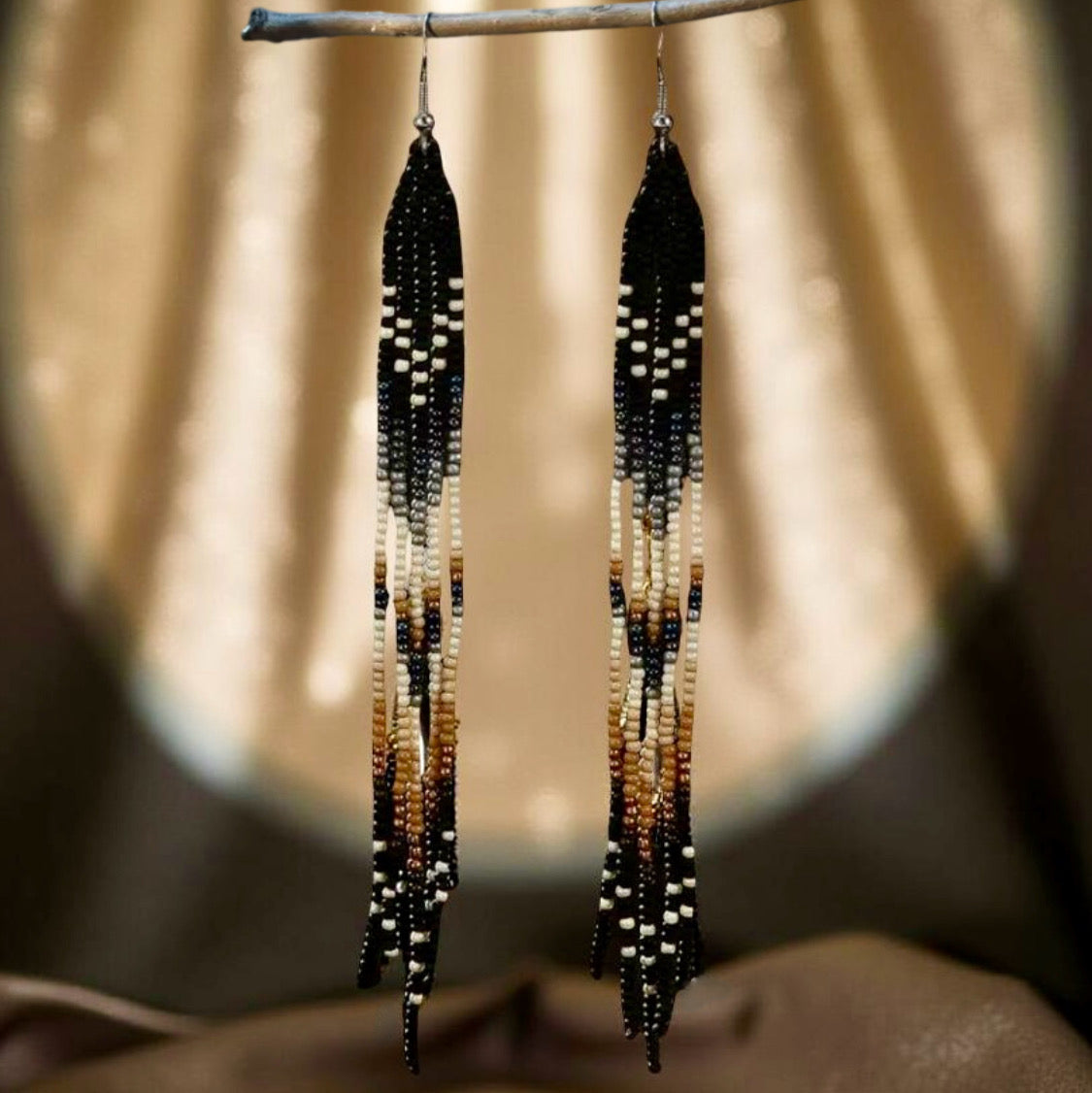 A pair of long handmade beaded earrings featuring a black, cream and tan colour pattern with a flowing fringe design, hanging against a soft spotlight background.