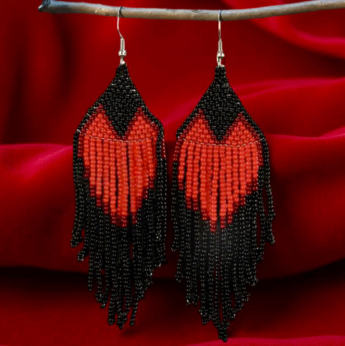 Handmade beaded earrings with a red heart design and long black fringe, displayed against a red velvet background on a wooden twig.