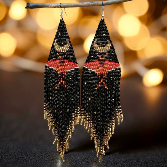 Long seed bead fringe earrings, featuring a red moth and gold crescent moon, in front of blurred bokeh lights