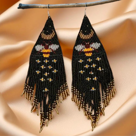 Handmade beaded earrings with a bee and crescent moon design, featuring long black and gold fringe, hanging from a twig on a satin background.