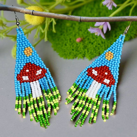 Handmade beaded earrings with a blue sky and mushroom design, featuring green and yellow fringe and hypoallergenic medical-grade 316L surgical steel lever backs.