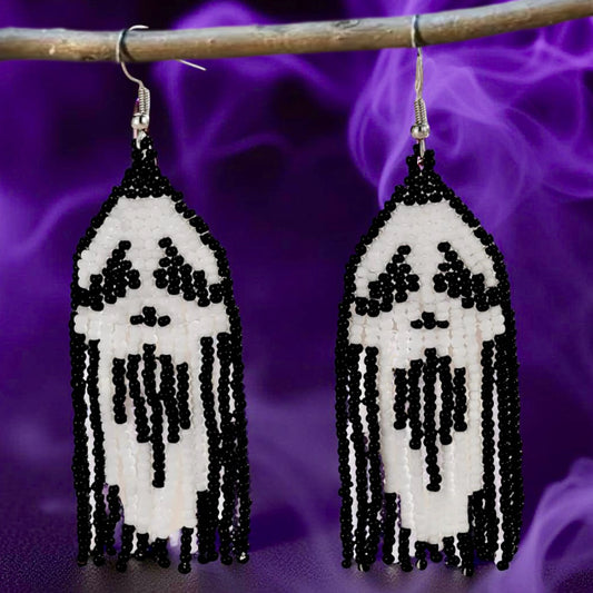 A pair of handmade beaded earrings featuring a black and white ghost face design in the fringe, hanging from hooks against a purple smokey background.