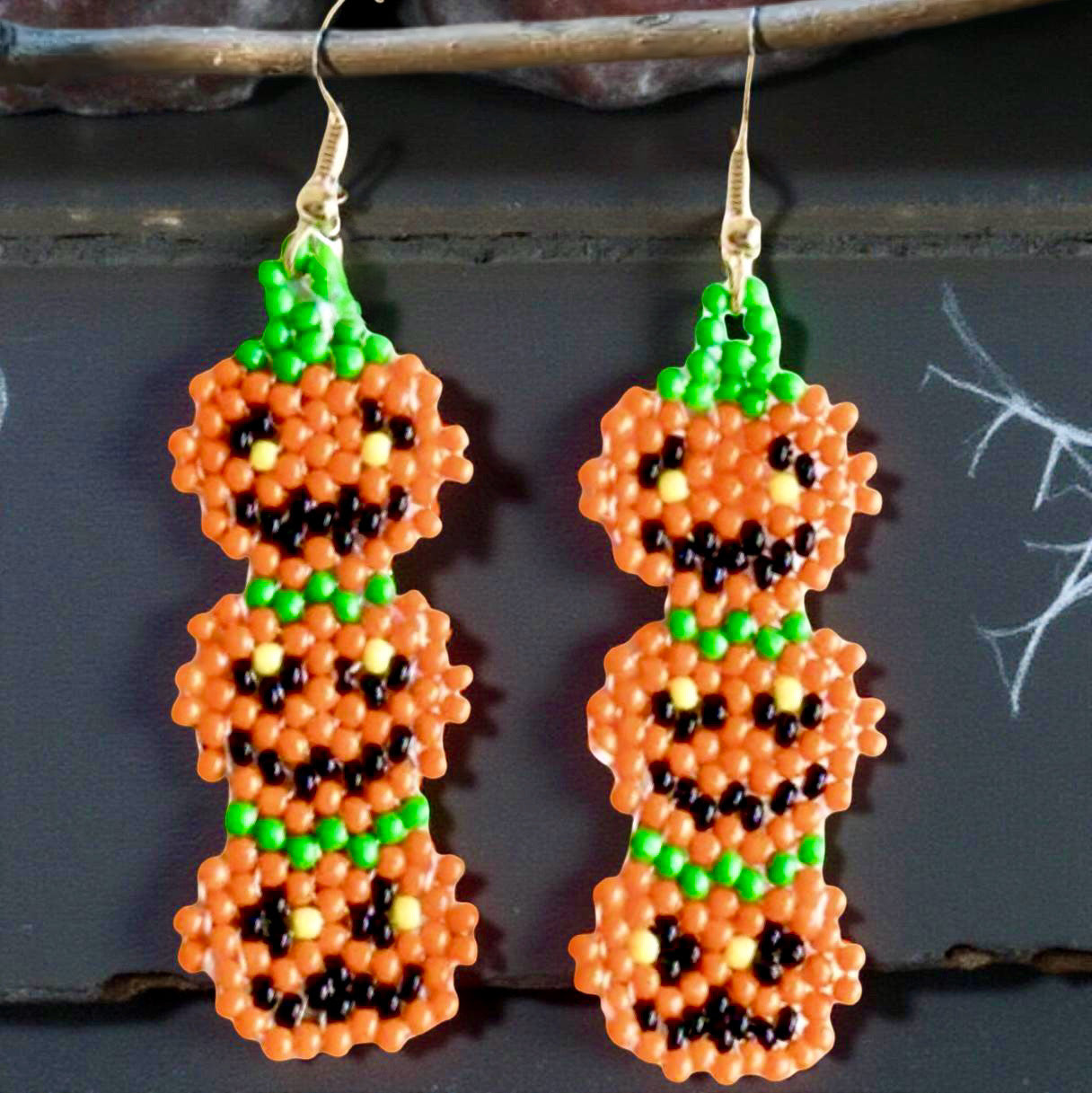 Handmade beaded earrings featuring a vertical stack of three smiling jack-o'-lantern pumpkins in orange, green, and black, perfect for Halloween-themed outfits. Black background