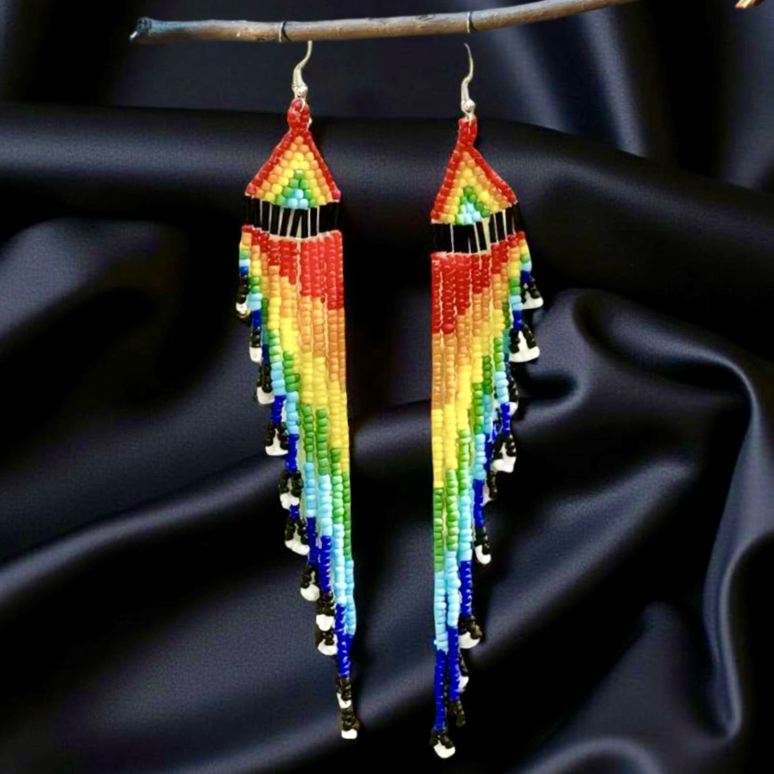 Long handmade beaded earrings with a vibrant rainbow fringe, displayed against a black satin background on a wooden hanger.