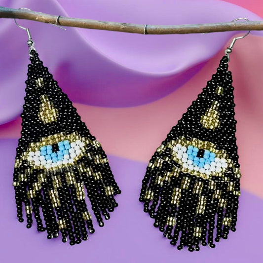 Handmade beaded earrings featuring a black triangle design with a central blue all-seeing eye and black and gold fringe, hanging on wire hooks. Pastel background