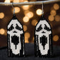 A pair of handmade beaded earrings featuring a black and white ghost face design in the fringe, hanging from hooks against a bokeh background.