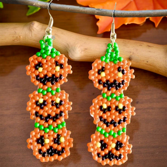 Handmade beaded earrings featuring a vertical stack of three smiling jack-o'-lantern pumpkins in orange, green, and black, perfect for Halloween-themed outfits.