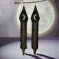 A pair of handmade beaded earrings hanging from a branch, featuring a crescent moon design and long black fringe, with a full moon softly glowing in the background.