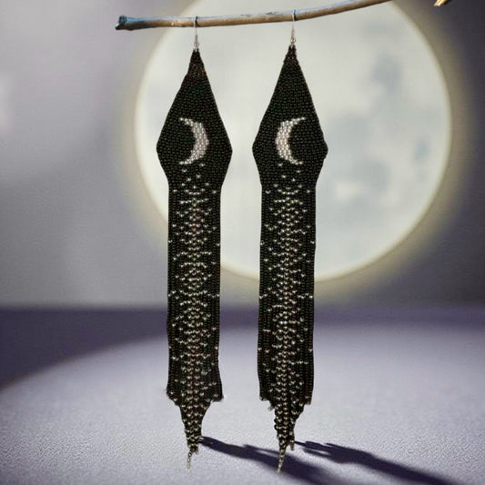 A pair of handmade beaded earrings hanging from a branch, featuring a crescent moon design and long black fringe, with a full moon softly glowing in the background.