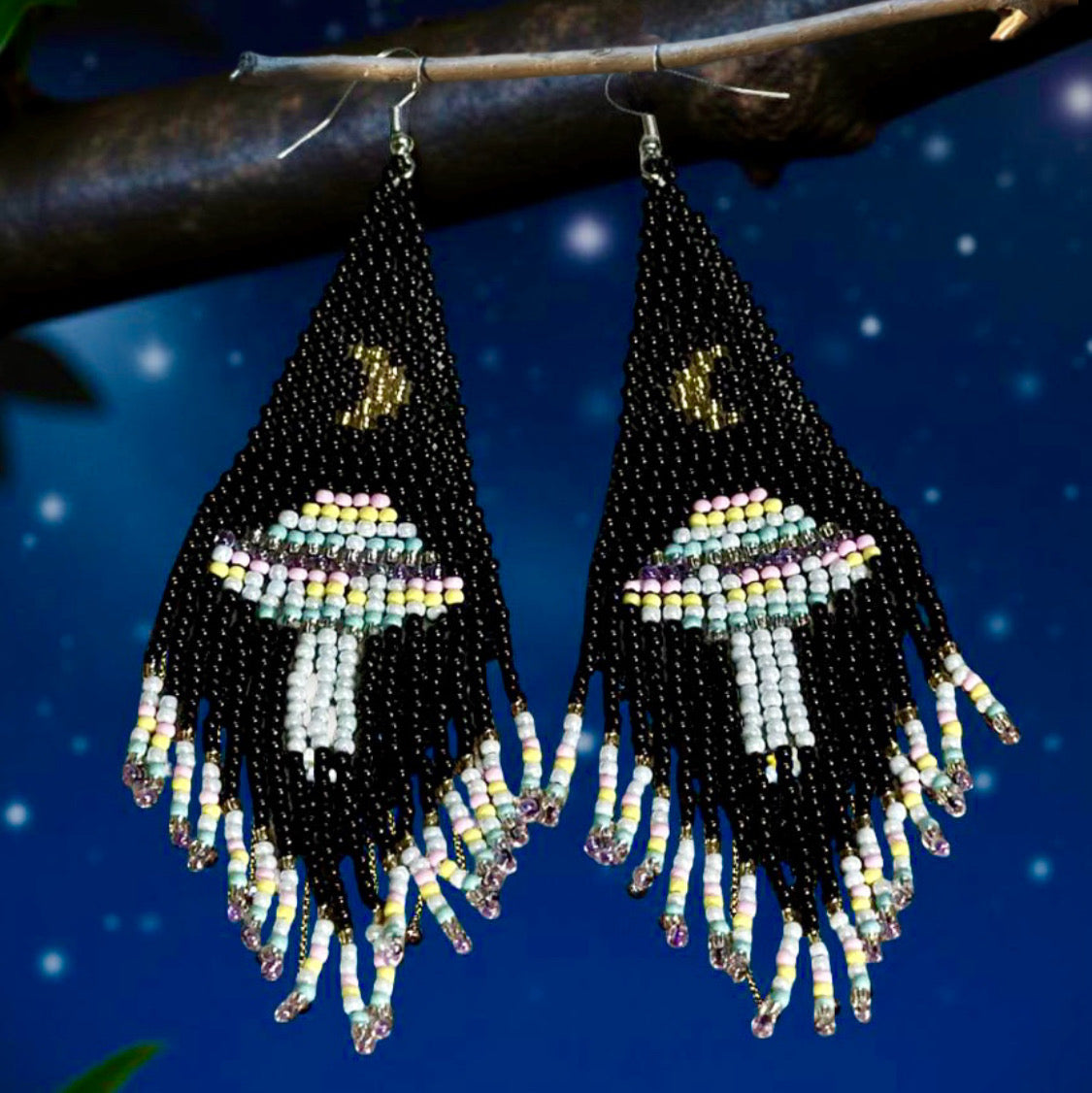 Handmade beaded earrings with black fringe, featuring a UFO and moon design. The earrings are mounted on surgical steel lever backs and show intricate bead detailing. Night sky background 