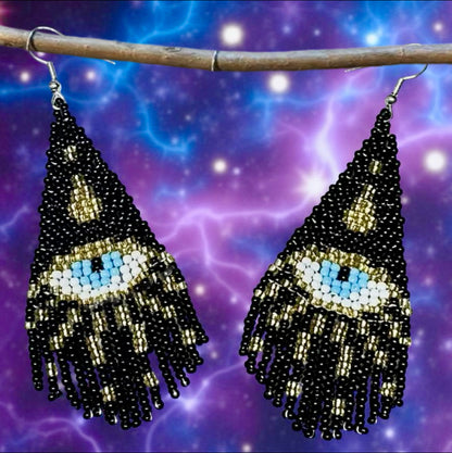 Handmade beaded earrings featuring a black triangle design with a central blue all-seeing eye and black and gold fringe, hanging on wire hooks.