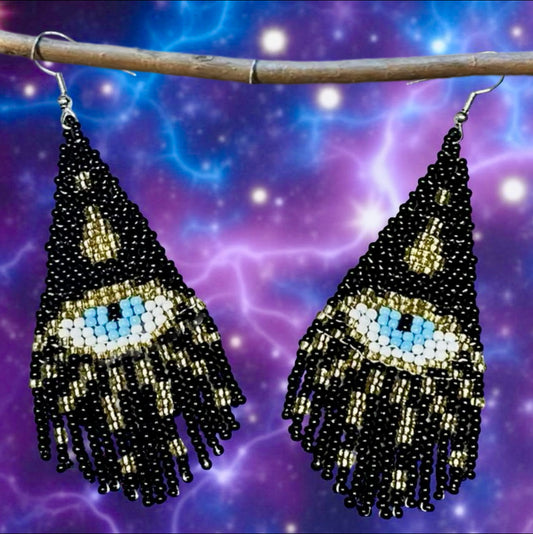 Handmade beaded earrings featuring a black triangle design with a central blue all-seeing eye and black and gold fringe, hanging on wire hooks.