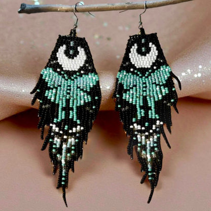 Handmade beaded earrings with a moon and turquoise butterfly design, featuring a black fringe. The earrings are hung from hypoallergenic 316L surgical steel lever backs. Shimmery fabric background 