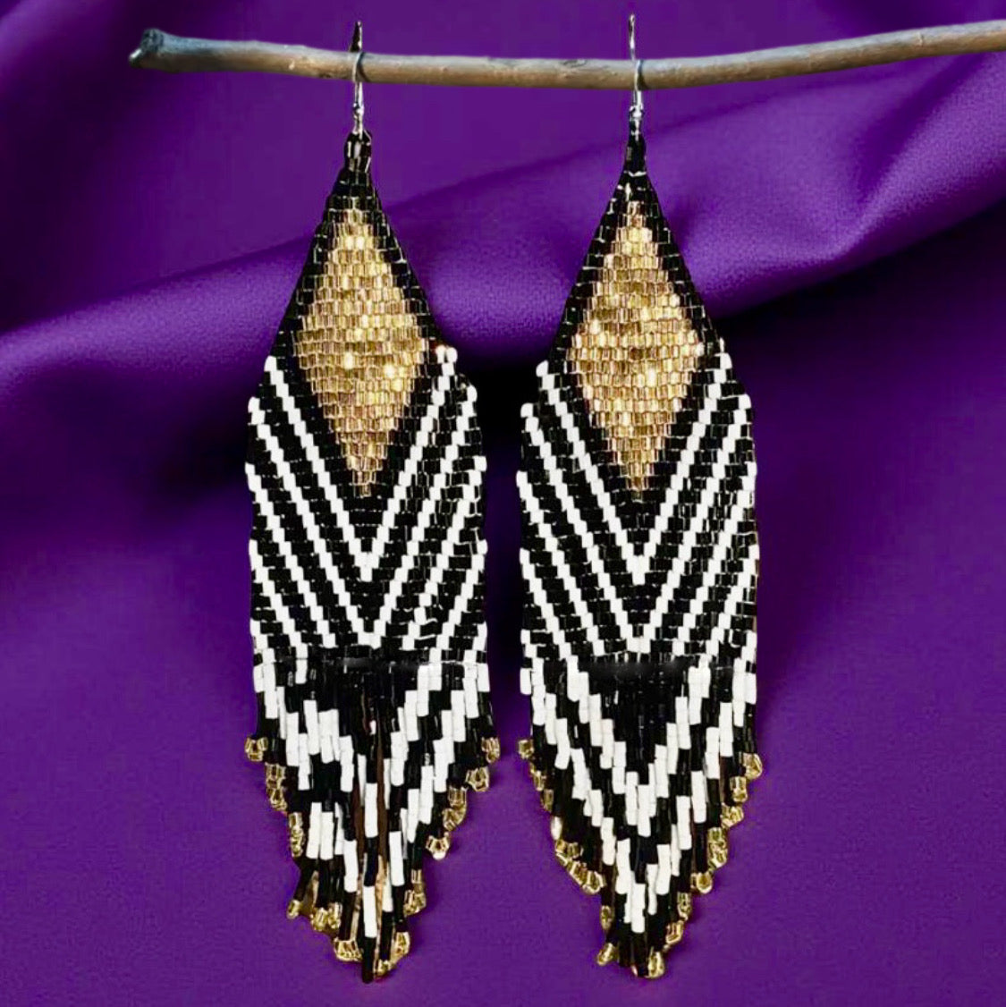 A pair of handmade beaded earrings with long black, gold, and white fringes hanging from hypoallergenic surgical steel lever backs, displayed against a purple fabric background.