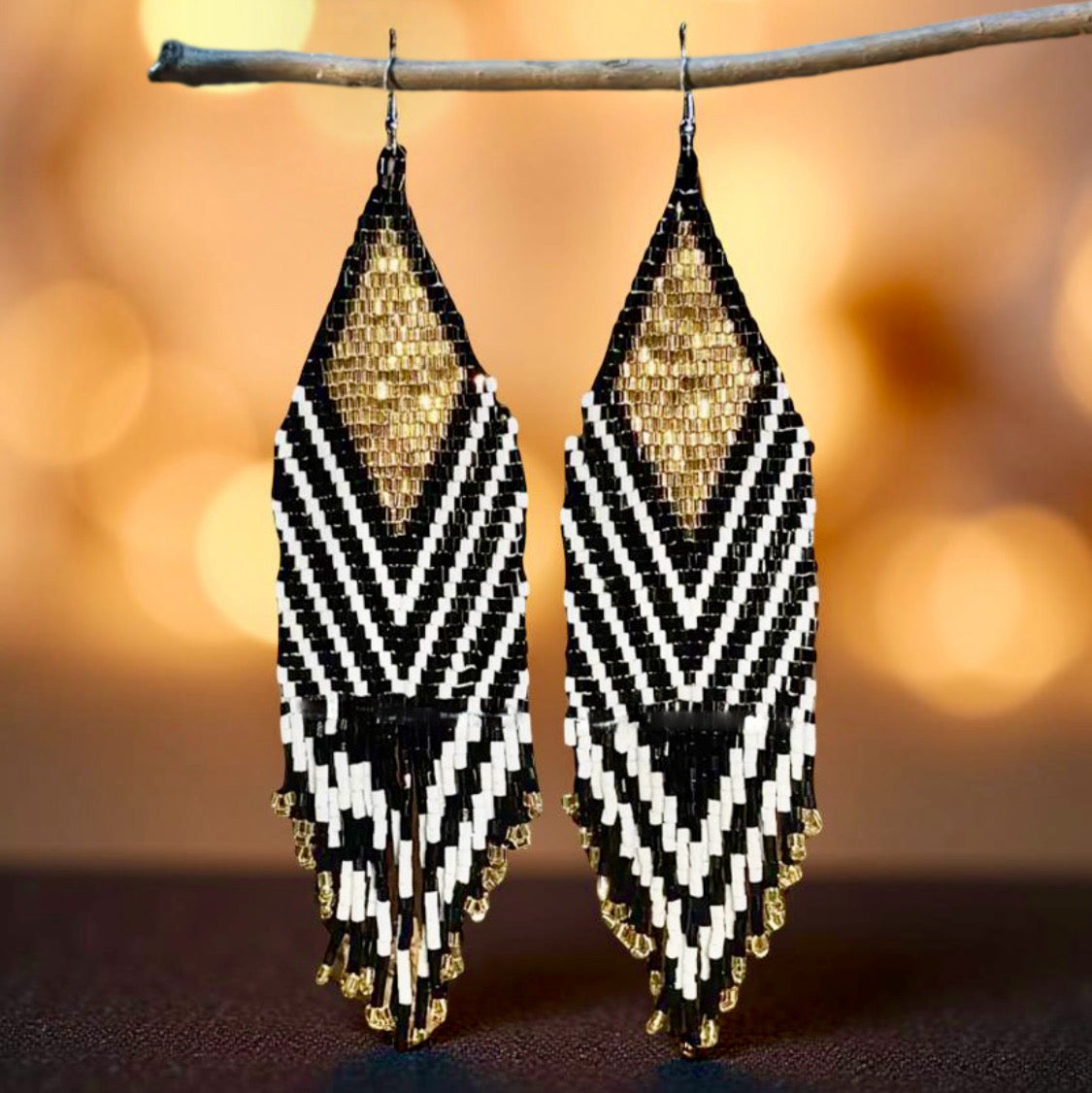 A pair of handmade beaded earrings with long black, gold, and white fringes hanging from hypoallergenic surgical steel lever backs, displayed against blurred bokeh lights