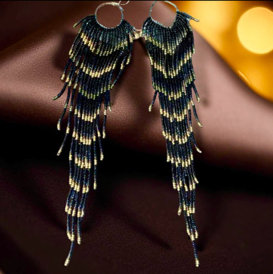 Handmade beaded earrings with extra-long black, green, and gold fringe displayed on a soft chocolate satin background.