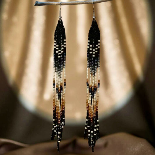 A pair of long handmade beaded earrings featuring a black, cream and tan colour pattern with a flowing fringe design, hanging against a soft spotlight background.