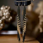 A pair of long handmade beaded earrings with a sleek black and gold fringe design, hanging from hooks against a blurred golden-toned background.