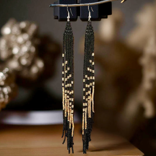 A pair of long handmade beaded earrings with a sleek black and gold fringe design, hanging from hooks against a blurred golden-toned background.