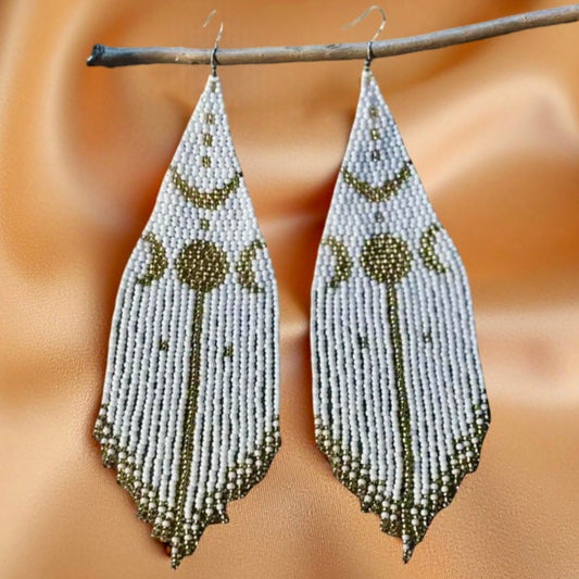 Handmade beaded earrings with a white and gold moon phase design, hanging from a twig against a soft satin background.
