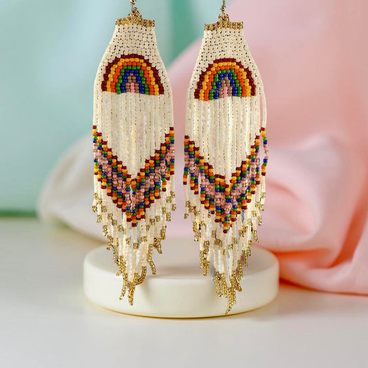 Handmade beaded earrings with a rainbow design at the top and long fringe, featuring colourful beads in red, yellow, green, blue, and purple. The earrings are finished with hypoallergenic 316L surgical steel lever backs. Pastel background 