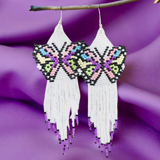 A pair of handmade beaded earrings featuring a multicoloured butterfly design with black accents and a long white fringe, displayed on a purple fabric background.