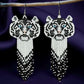 Handmade beaded earrings featuring a white tiger face with blue eyes and a long black, white, and grey beaded fringe, hanging from surgical steel lever backs. Dark satin background 