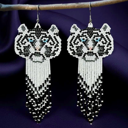 Handmade beaded earrings featuring a white tiger face with blue eyes and a long black, white, and grey beaded fringe, hanging from surgical steel lever backs. Dark satin background 