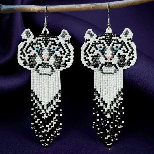 Handmade beaded earrings featuring a white tiger face with blue eyes and a long black, white, and grey beaded fringe, hanging from surgical steel lever backs. Dark satin background 
