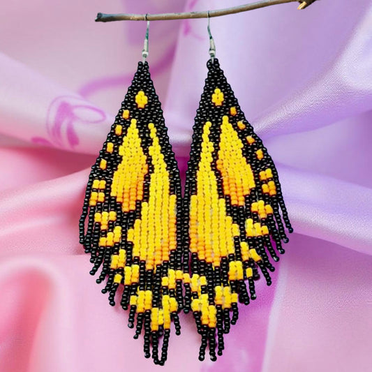 Handmade beaded earrings designed to resemble yellow butterfly wings with black accents, displayed against a soft pink and purple background.