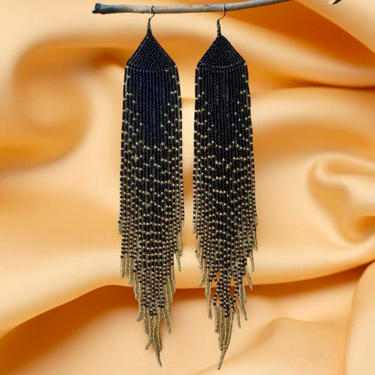 Handmade beaded earrings designed to mimic long feathers, featuring a black and gold cascading bead pattern with hypoallergenic 316L surgical steel lever backs, against a satin background 