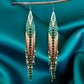 Handmade beaded earrings with long fringe, featuring a mix of teal, gold, black, and soft pink colors, and hypoallergenic 316L surgical steel lever backs.