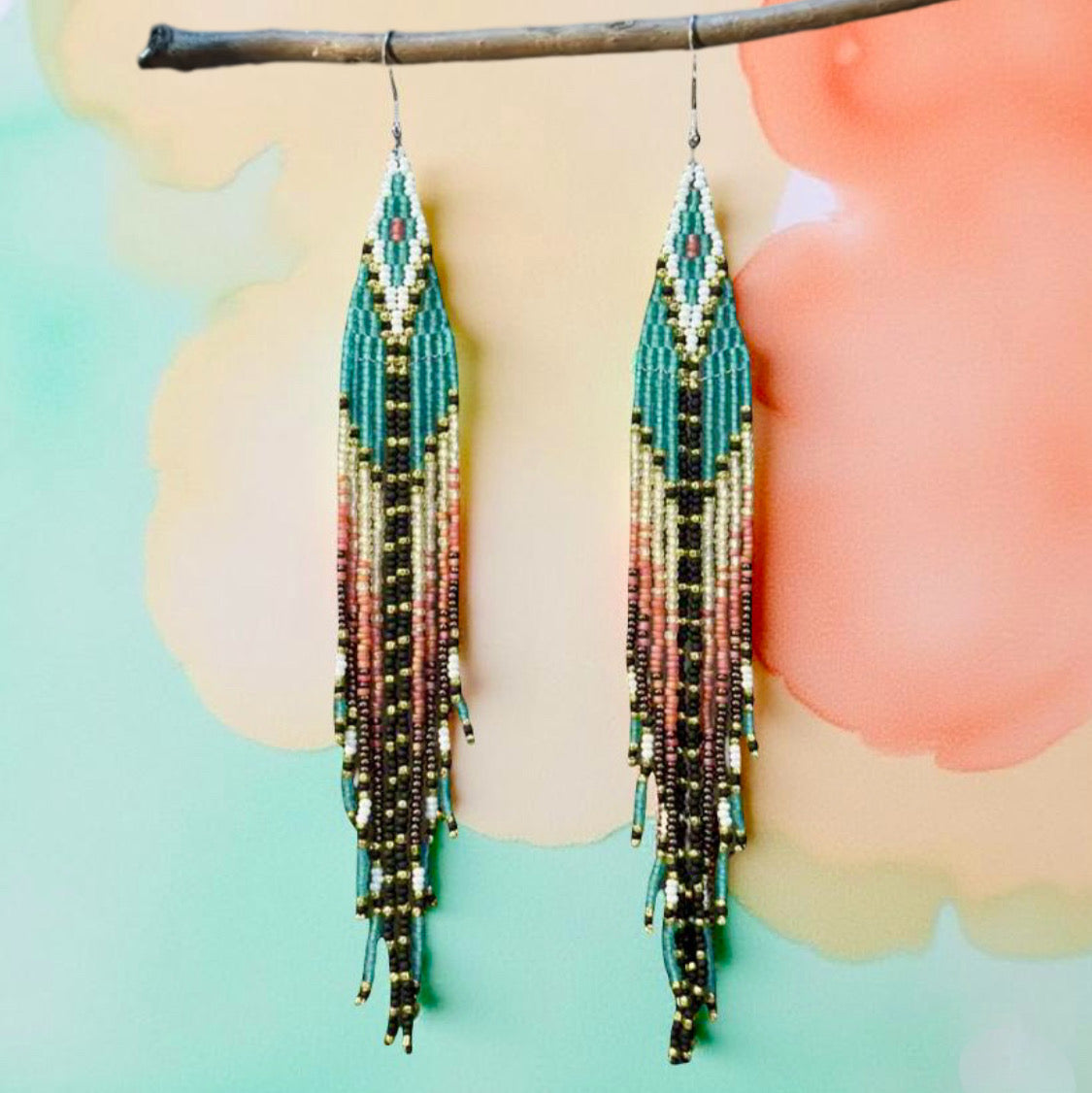Handmade beaded earrings with long fringe, featuring a mix of teal, gold, black, and soft pink colors, and hypoallergenic 316L surgical steel lever backs, against a watercolour background 