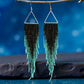 A pair of handmade beaded earrings featuring a triangular top and long black and teal fringe, hanging from hypoallergenic 316L surgical steel lever back hooks in front of a blue background 