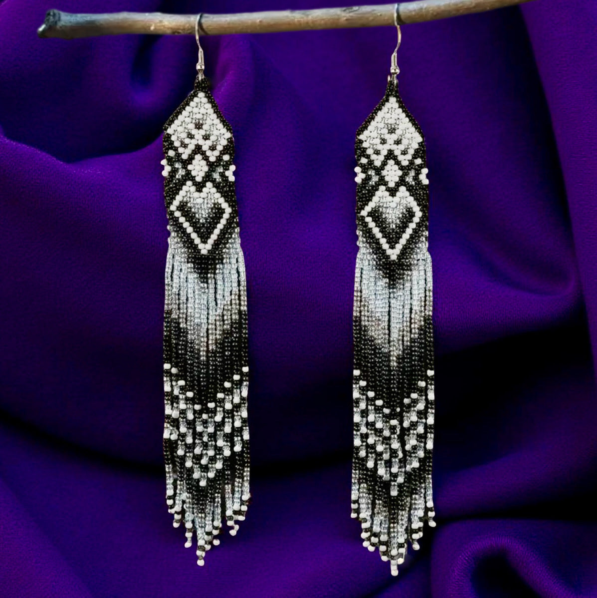 Handmade extra long black and white beaded fringe earrings with geometric diamond patterns, hanging on a wooden stick in front of a purple fabric background.