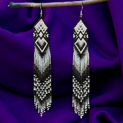 Handmade extra long black and white beaded fringe earrings with geometric diamond patterns, hanging on a wooden stick in front of a purple fabric background.