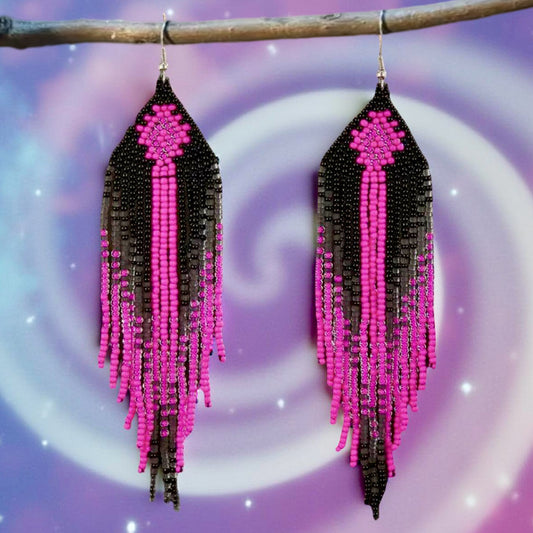 A pair of handmade beaded earrings featuring long pink and black fringe, with a diamond pattern at the top, hanging from surgical steel lever backs against a dreamy purple background.