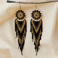 Handmade beaded earrings with a dream catcher-inspired design, featuring black, gold, and brown beads in a geometric pattern with a fringe. Hypoallergenic 316L surgical steel lever backs for comfort.