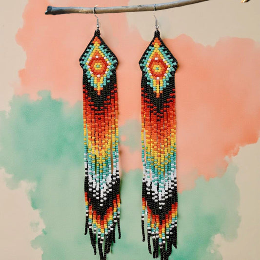 Handmade beaded earrings featuring a long fringe design with a vibrant colour palette of orange, red, yellow, turquoise, black, and white. The earrings have hypoallergenic 316L surgical steel lever backs. Watercolour background 