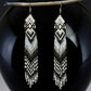 Handmade extra long black and white beaded fringe earrings with geometric diamond patterns, hanging on a wooden stick in front of a shiny black ball background.