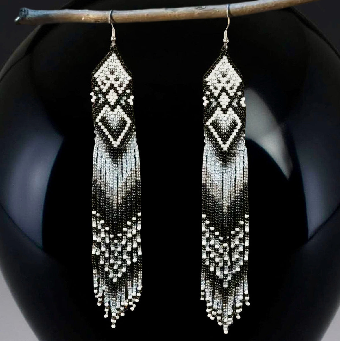 Handmade extra long black and white beaded fringe earrings with geometric diamond patterns, hanging on a wooden stick in front of a shiny black ball background.