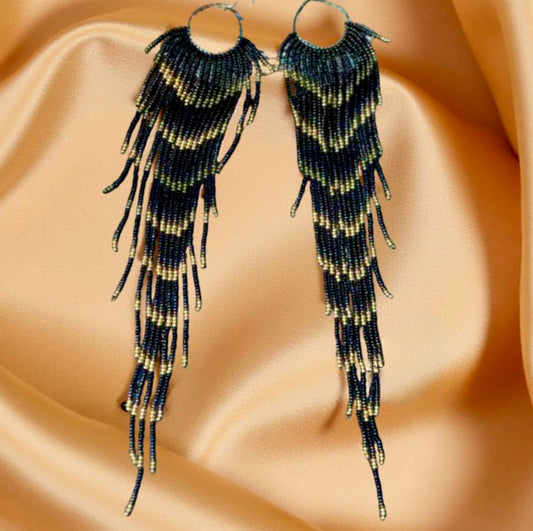 Handmade beaded earrings with extra-long black and gold fringe displayed on a soft beige satin background.